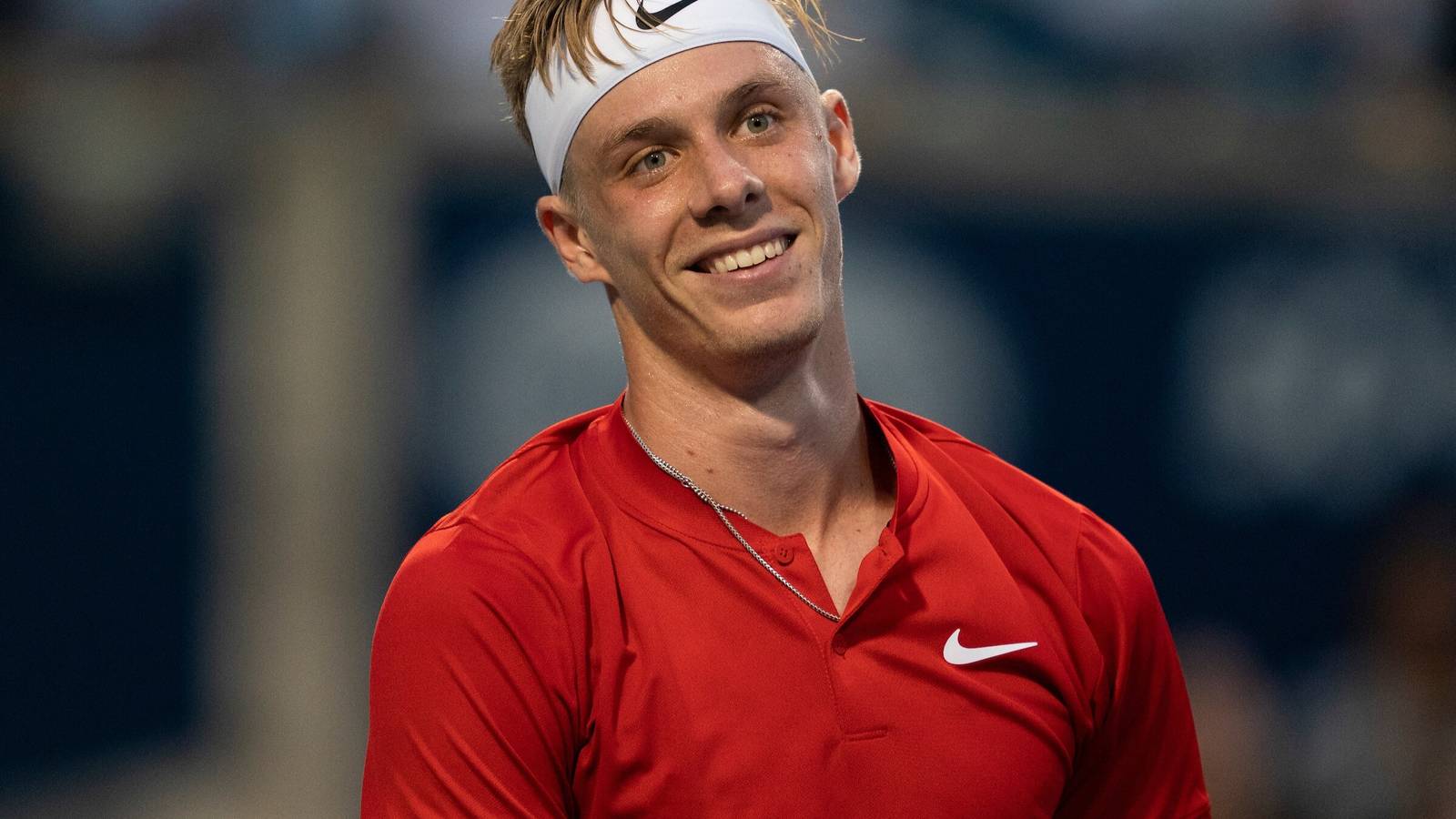 Canadian tennis player Denis Shapovalov wearing a white headband and red shirt smiling