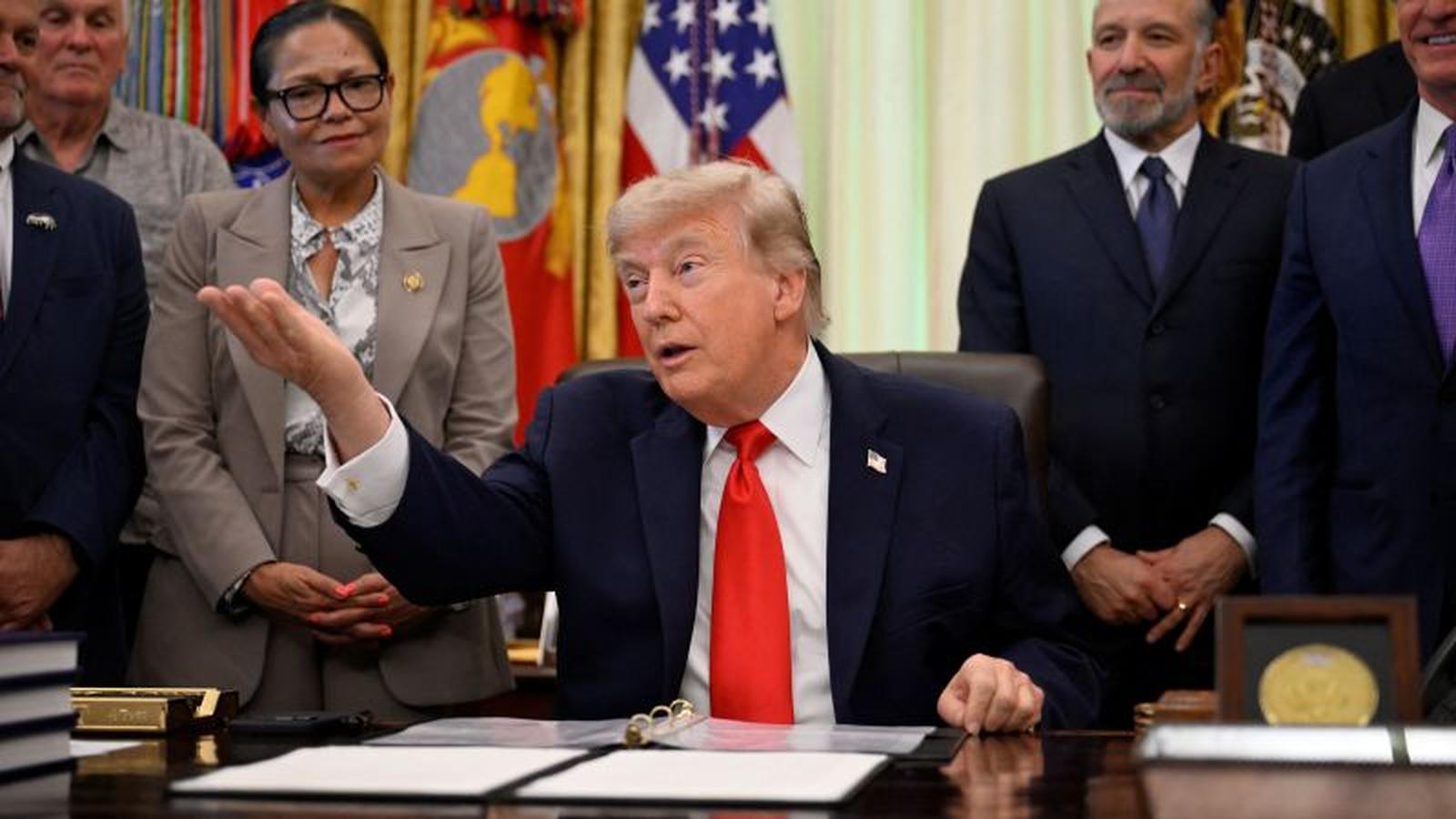 Donald Trump sitting at the Resolute Desk gesturing while surrounded by officials