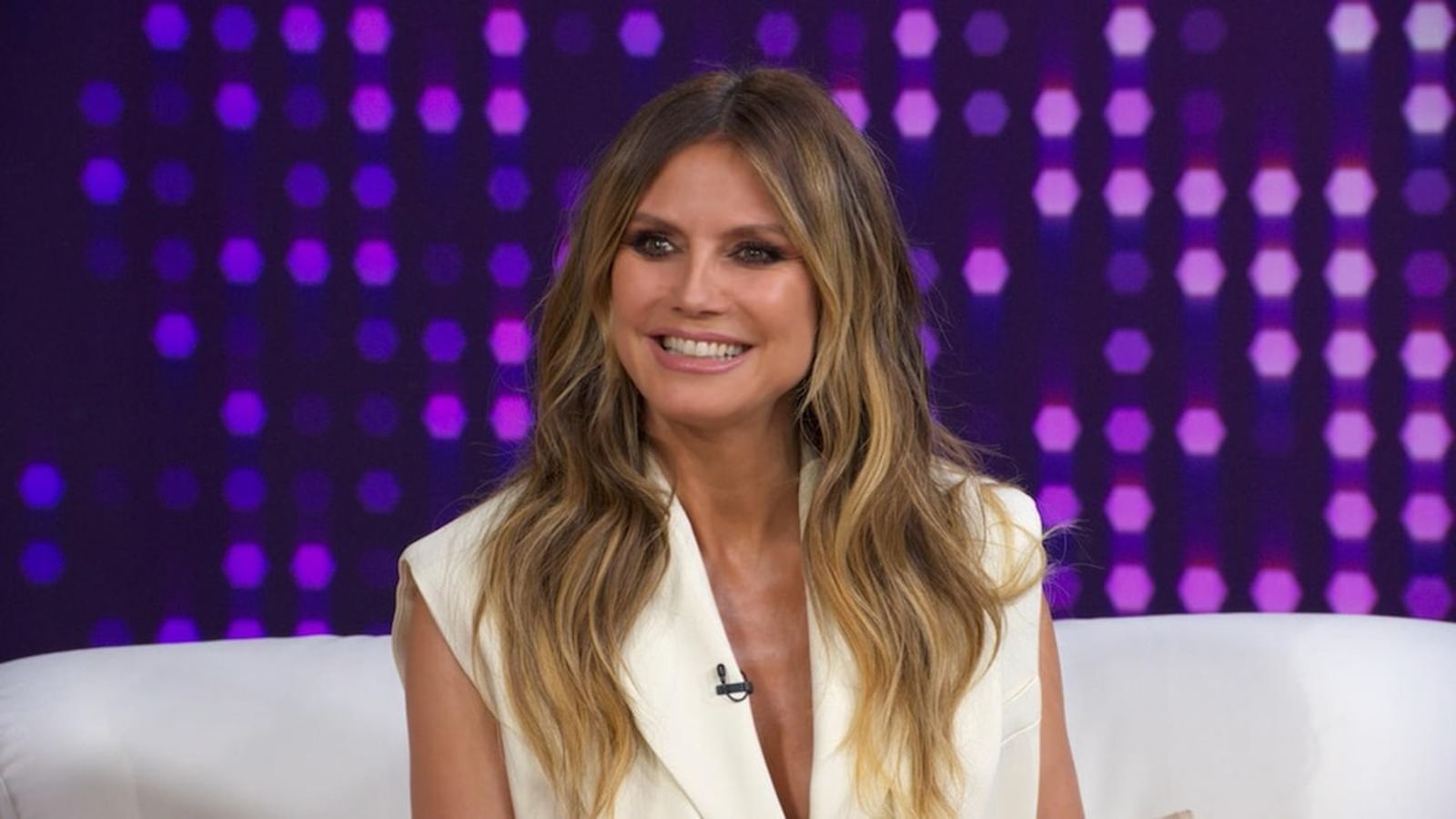 Heidi Klum smiling while wearing a white blazer in a television studio setting