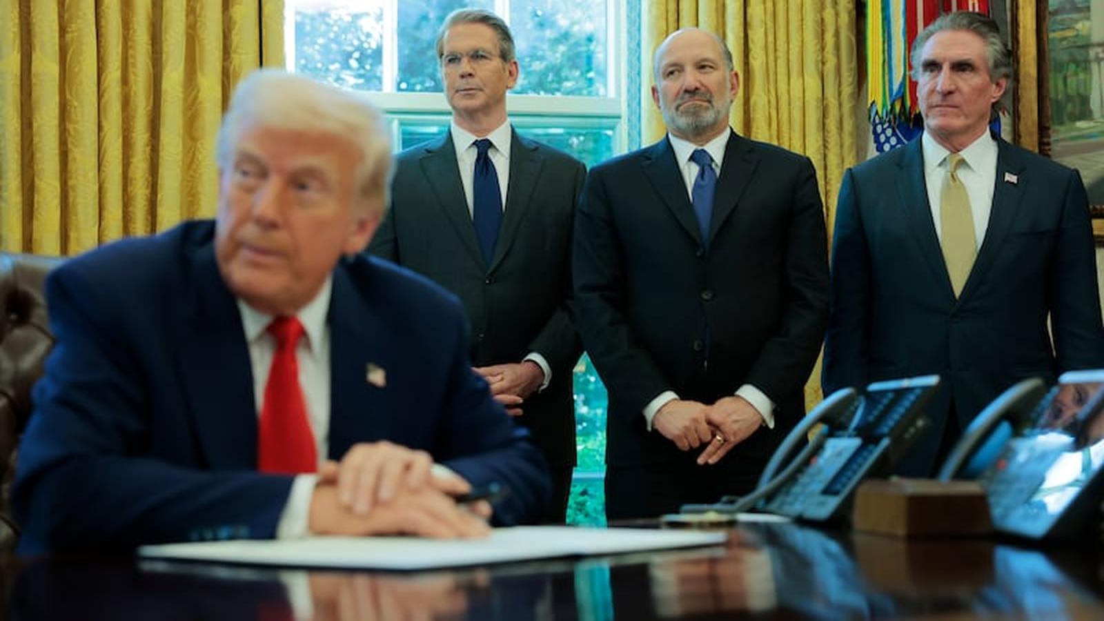 Donald Trump sits at a desk with Howard Lutnick and Scott Bessent standing behind