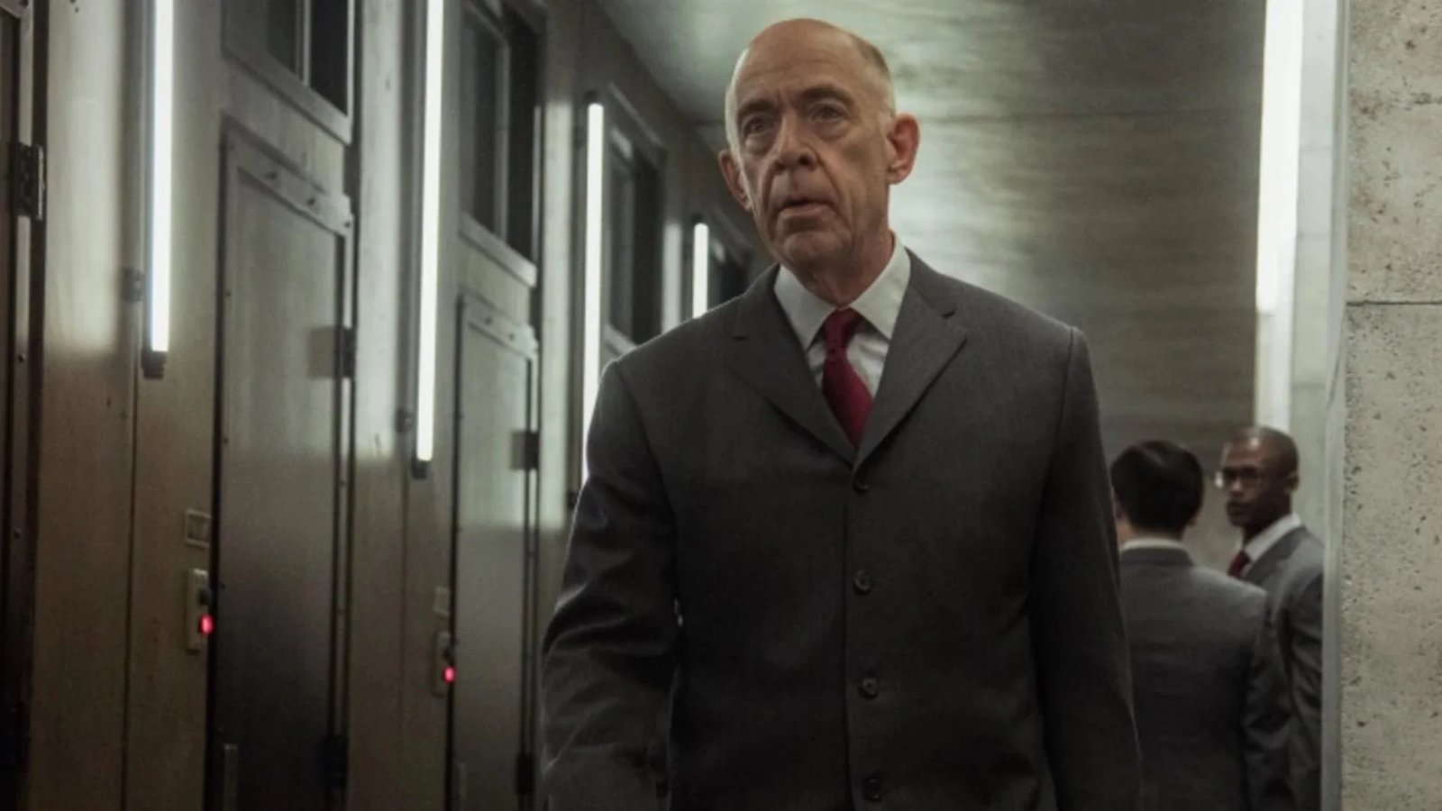 Actor J.K. Simmons wearing a grey suit and red tie walking down hallway