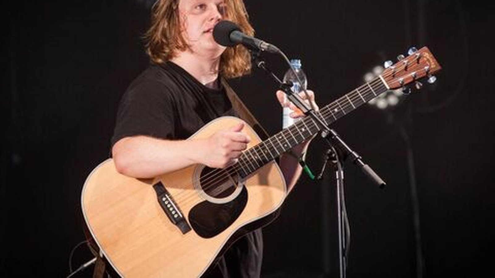 Lewis Capaldi singing and playing an acoustic guitar on a dark stage