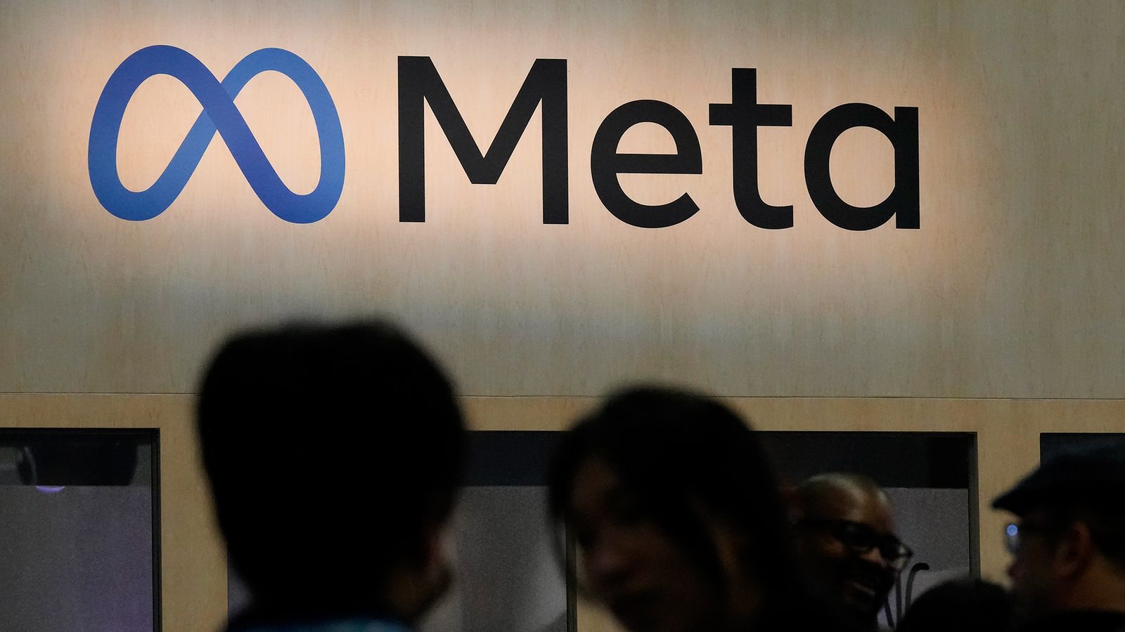 The Meta logo displayed on a wall with blurred silhouettes of people in foreground