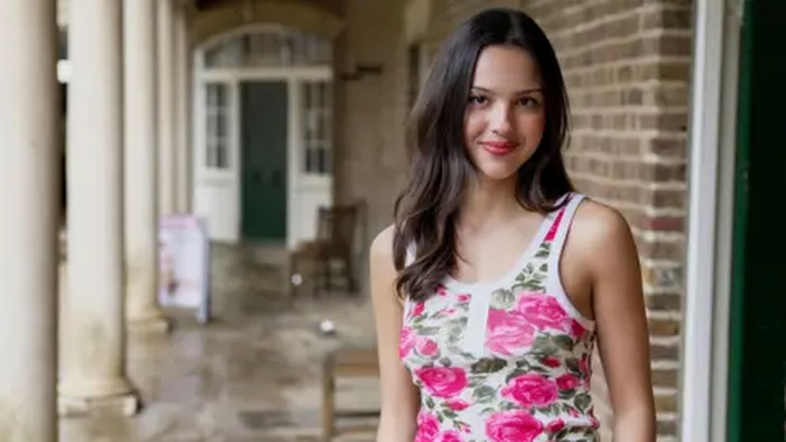 Olivia Rodrigo smiling while wearing a floral tank top in a hallway