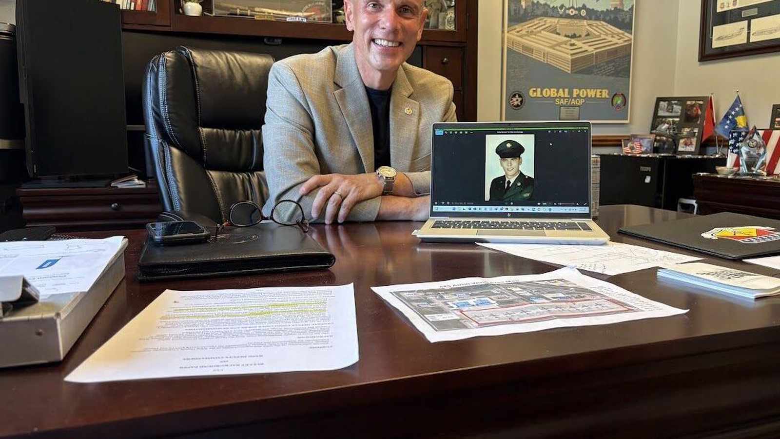 A smiling Pentagon official sits at a desk with a laptop displaying a military portrait
