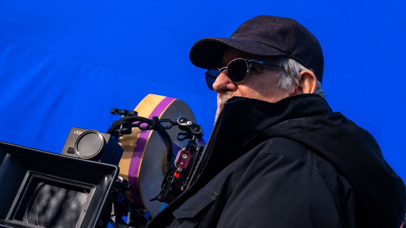 Steven Spielberg wearing a cap and sunglasses while operating a professional film camera