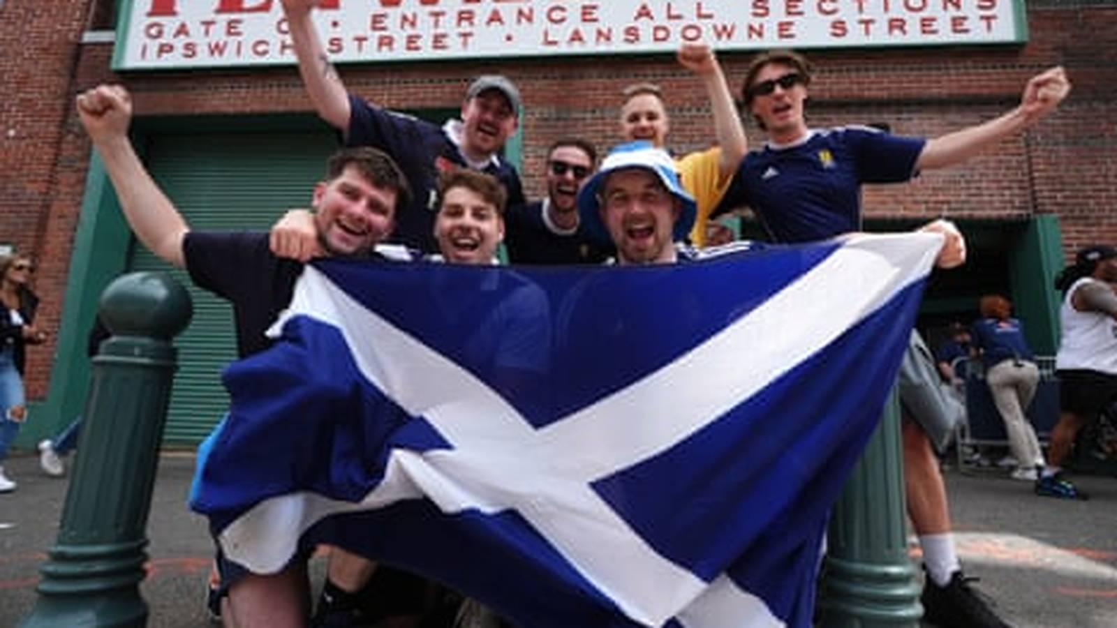 A group of cheering Scottish football fans holding a large Scottish flag outside Fenway Park