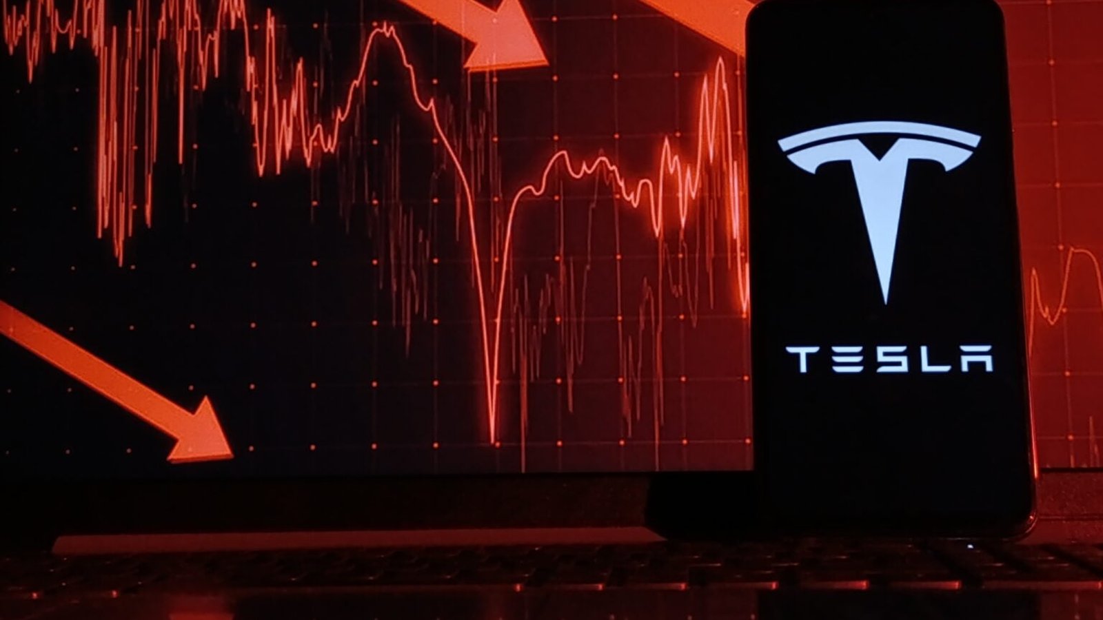 A smartphone displaying the Tesla logo in front of a red stock market chart