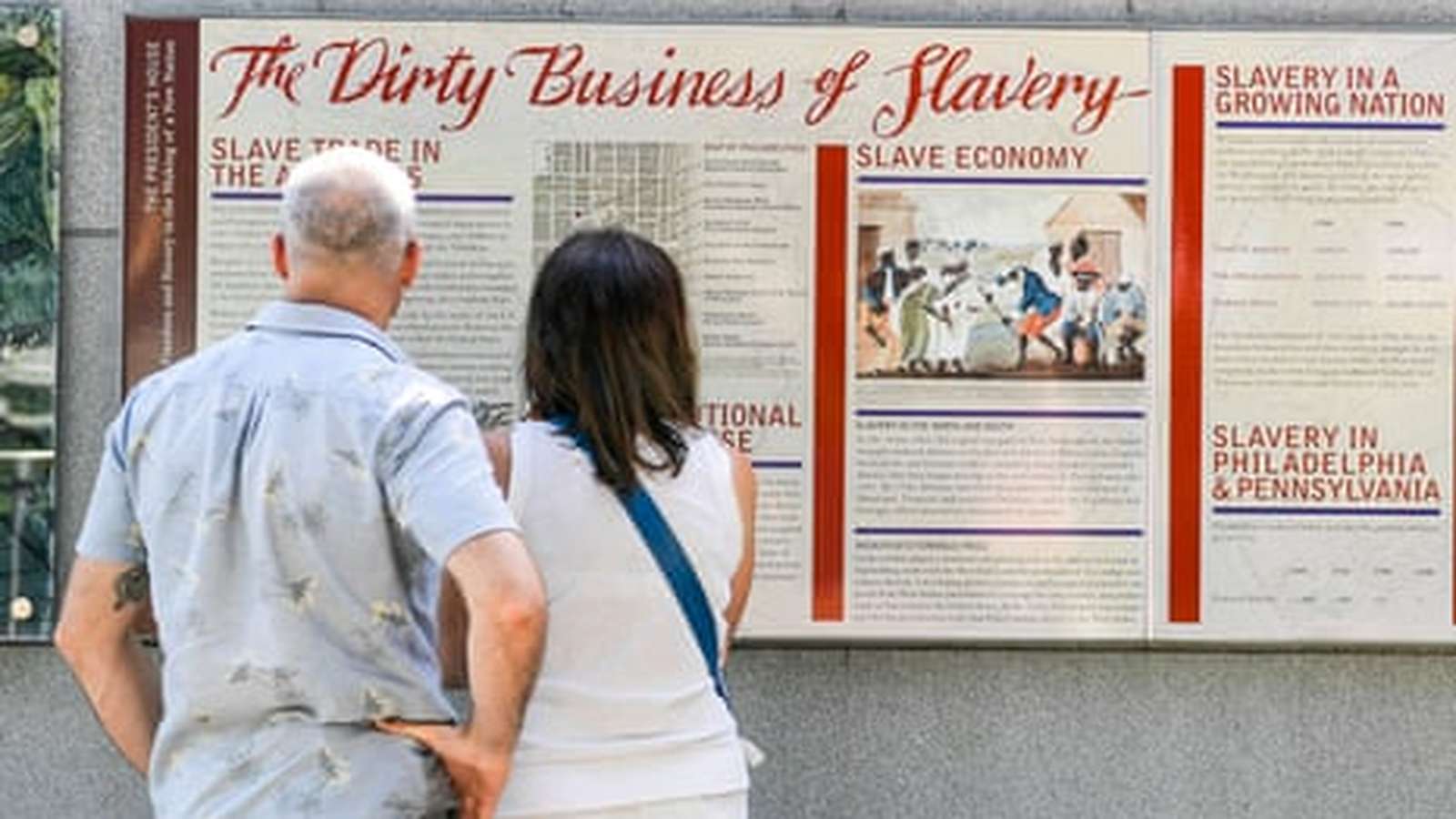 Two people standing and reading a historical information panel titled The Dirty Business of Slavery