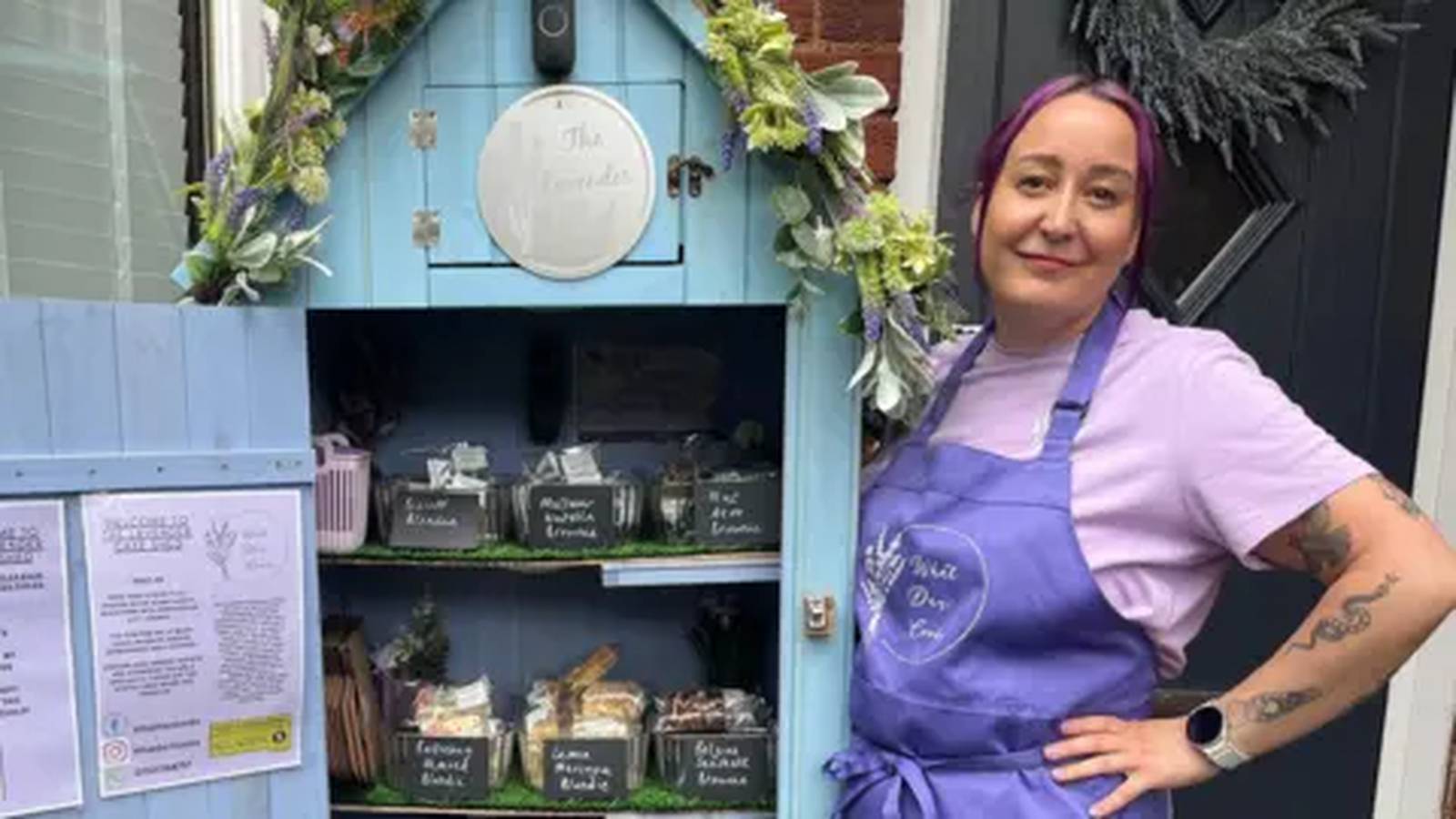 A woman wearing a purple apron stands beside a small blue honesty box cake shed