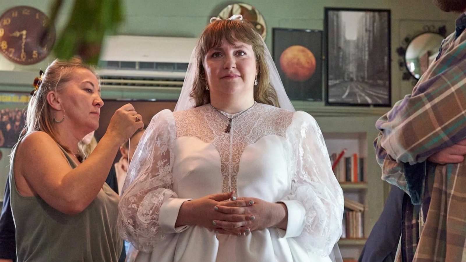 Lena Dunham wearing a white lace wedding dress while being assisted by a stylist
