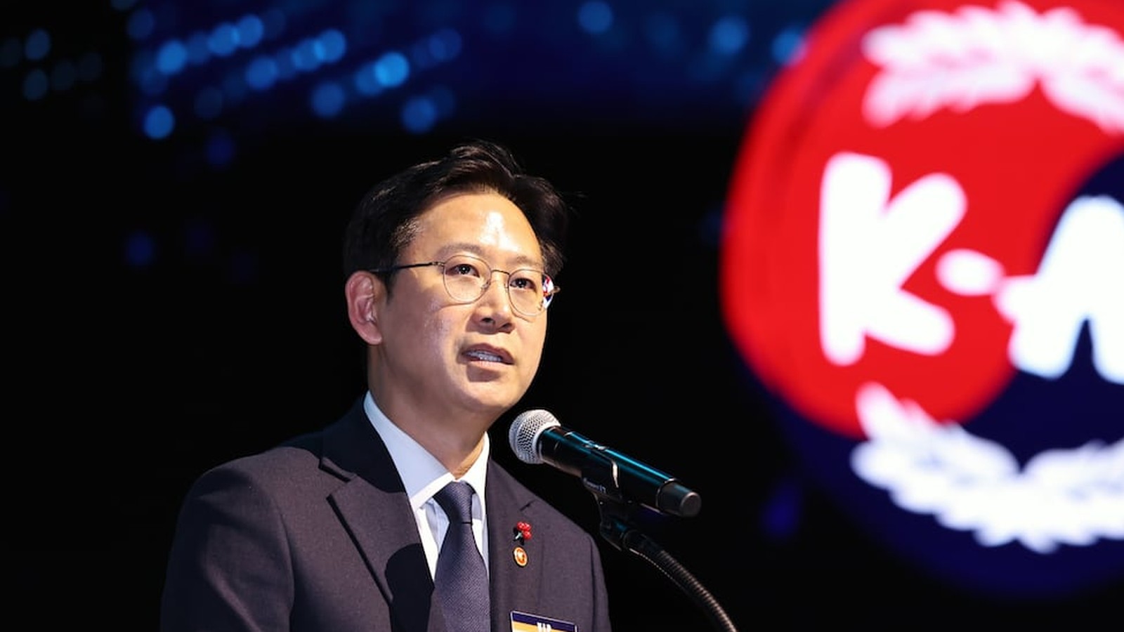 Bae Kyung-hoon speaking at a podium with a large K-AI logo in the background