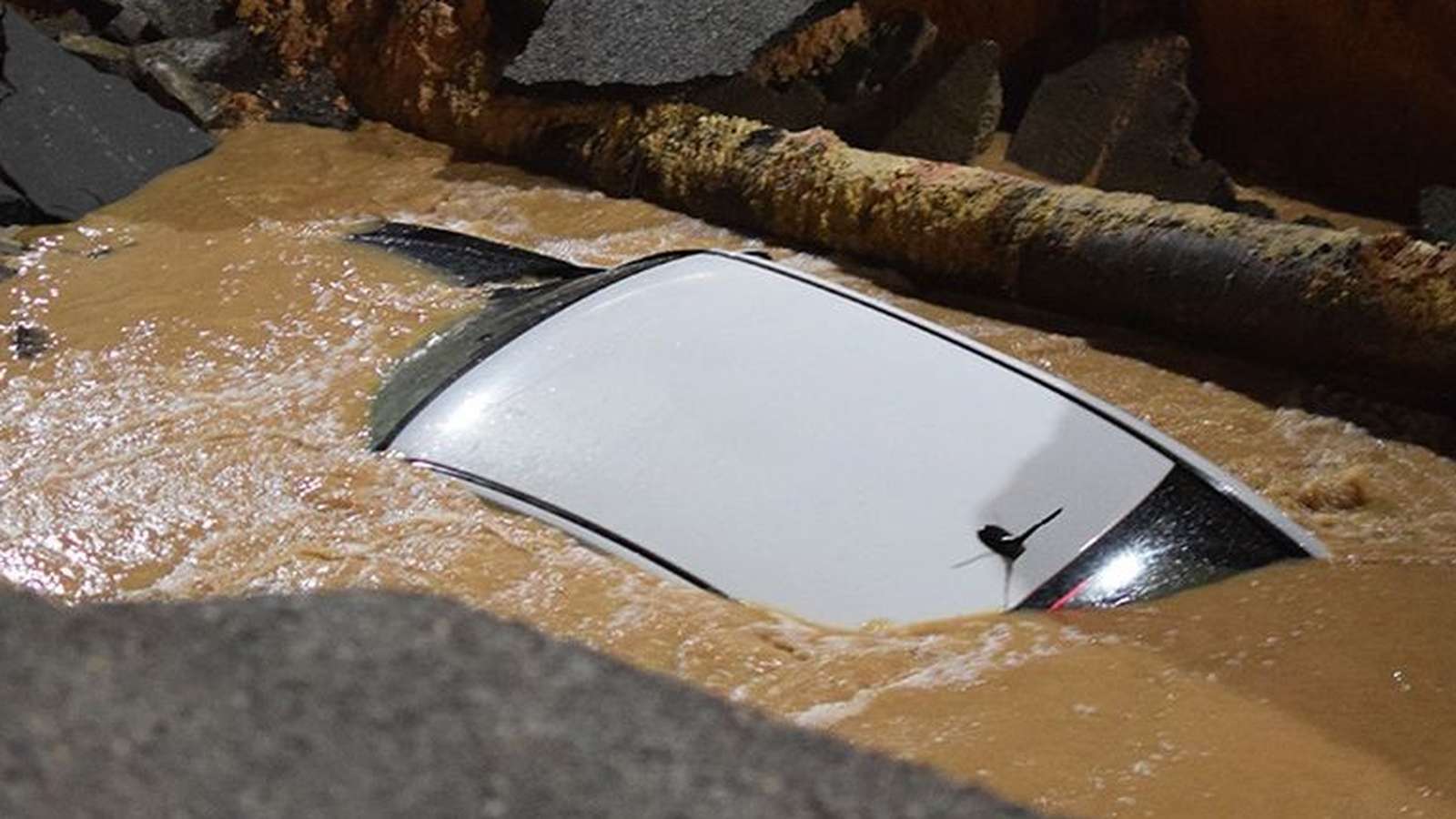 A white car partially submerged in muddy water inside a large urban sinkhole
