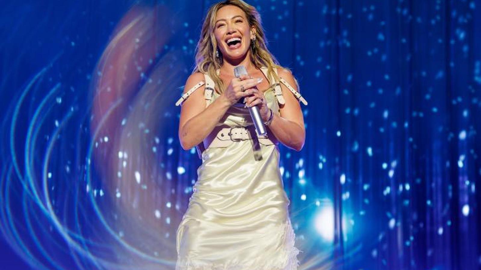 Hilary Duff smiling while singing into a microphone on a blue stage