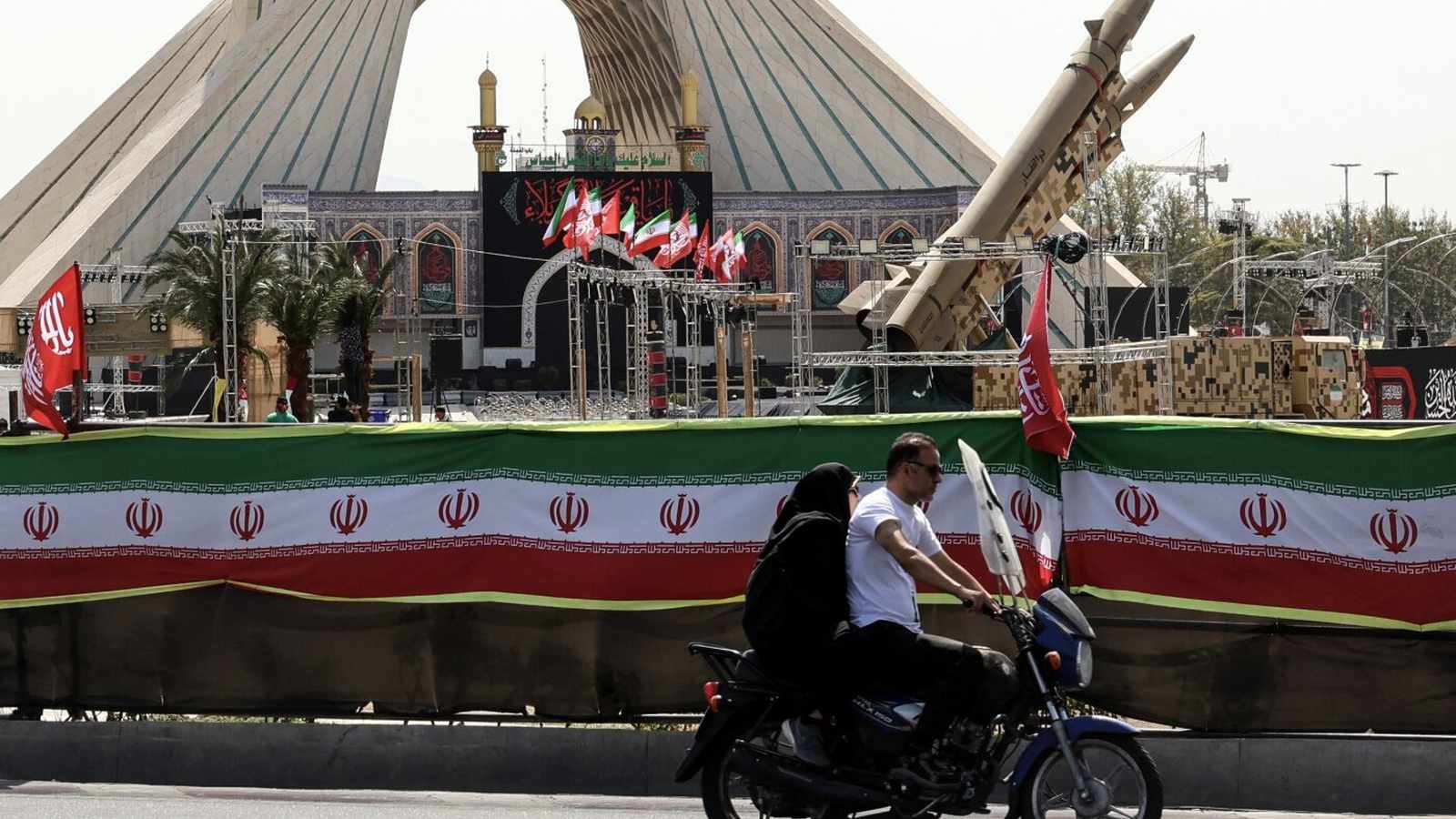 A couple riding a motorcycle past an Iranian military display featuring a missile