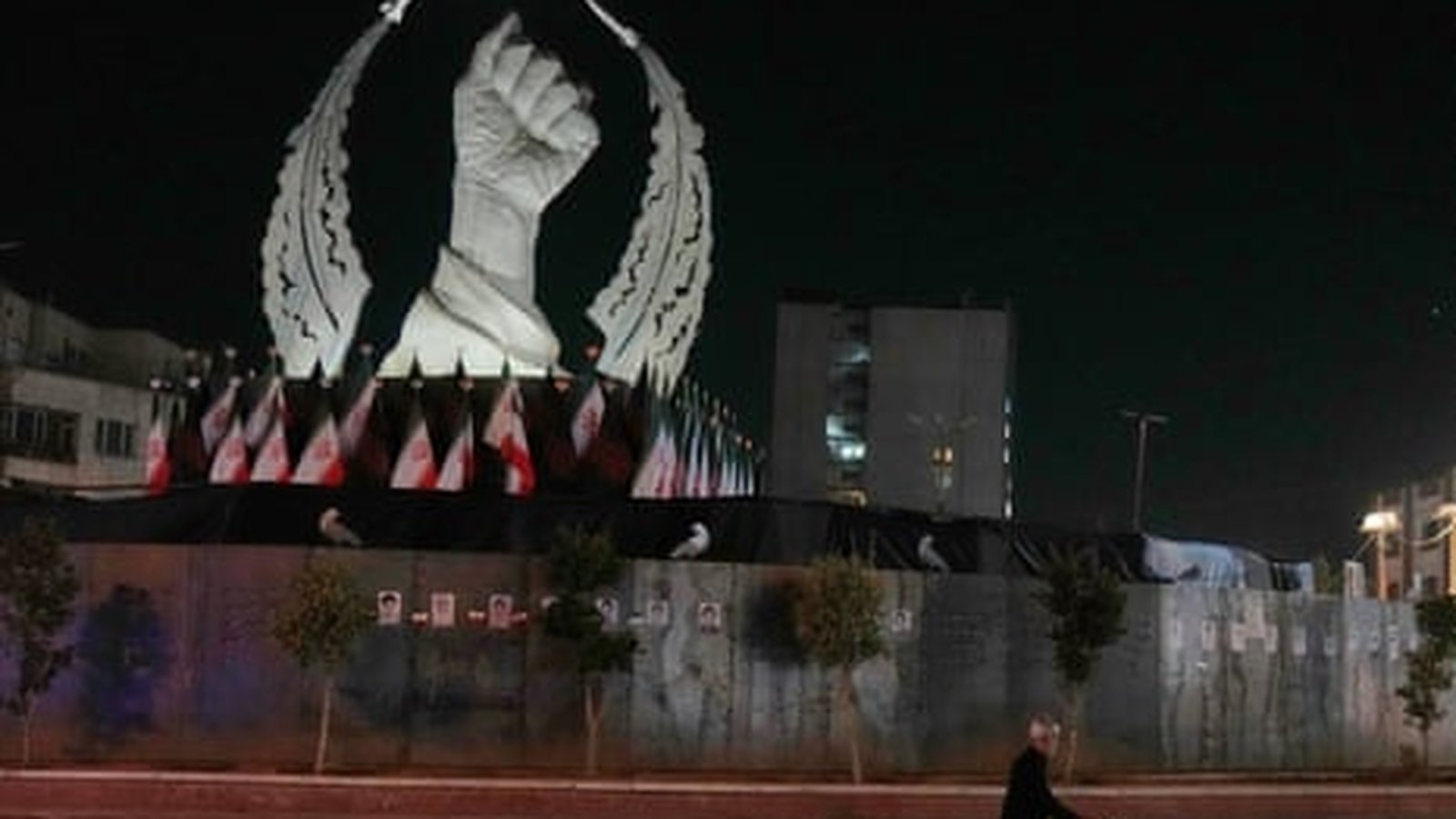 A large white fist monument surrounded by Iranian flags at night in Tehran