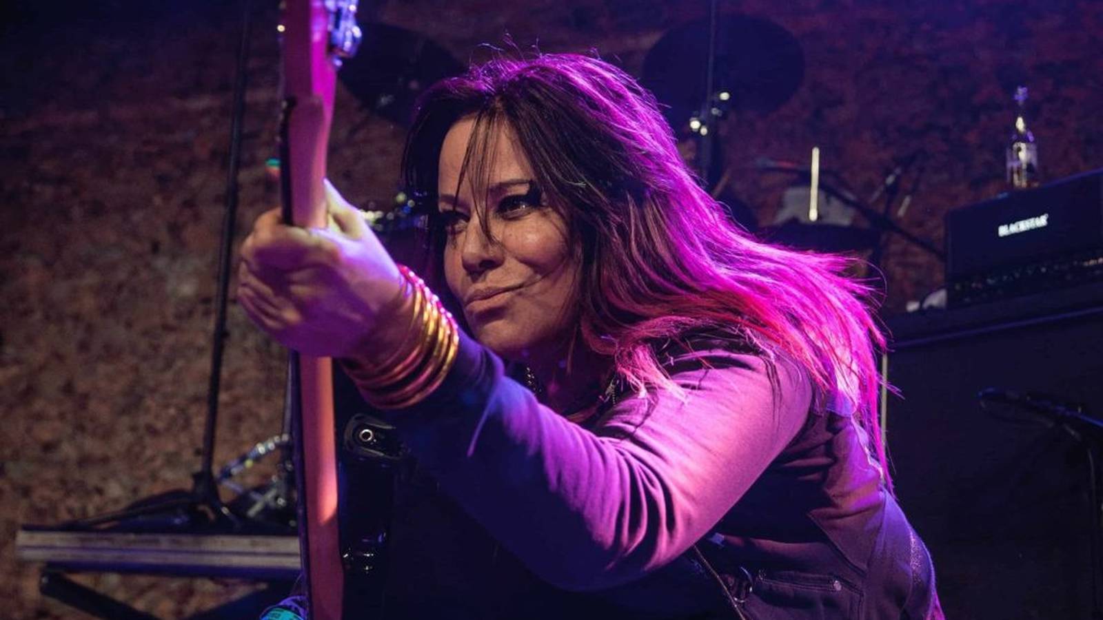 Jennifer Finch playing bass guitar on stage under purple stage lighting during concert