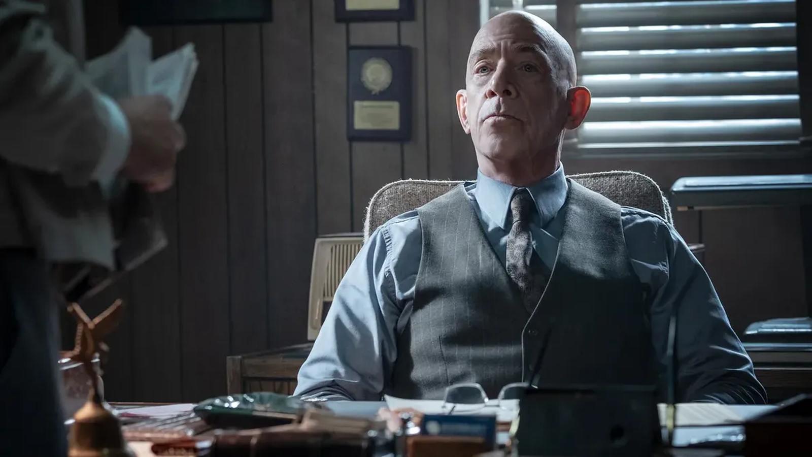 Actor JK Simmons sitting at a desk in a suit and vest