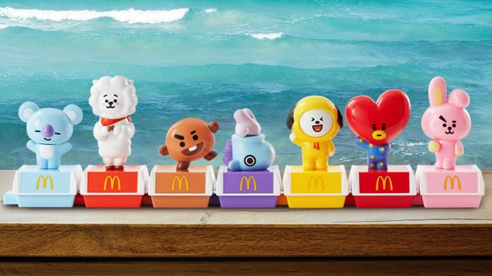 Seven colorful BT21 character figurines standing on McDonalds Happy Meal boxes against a beach backg