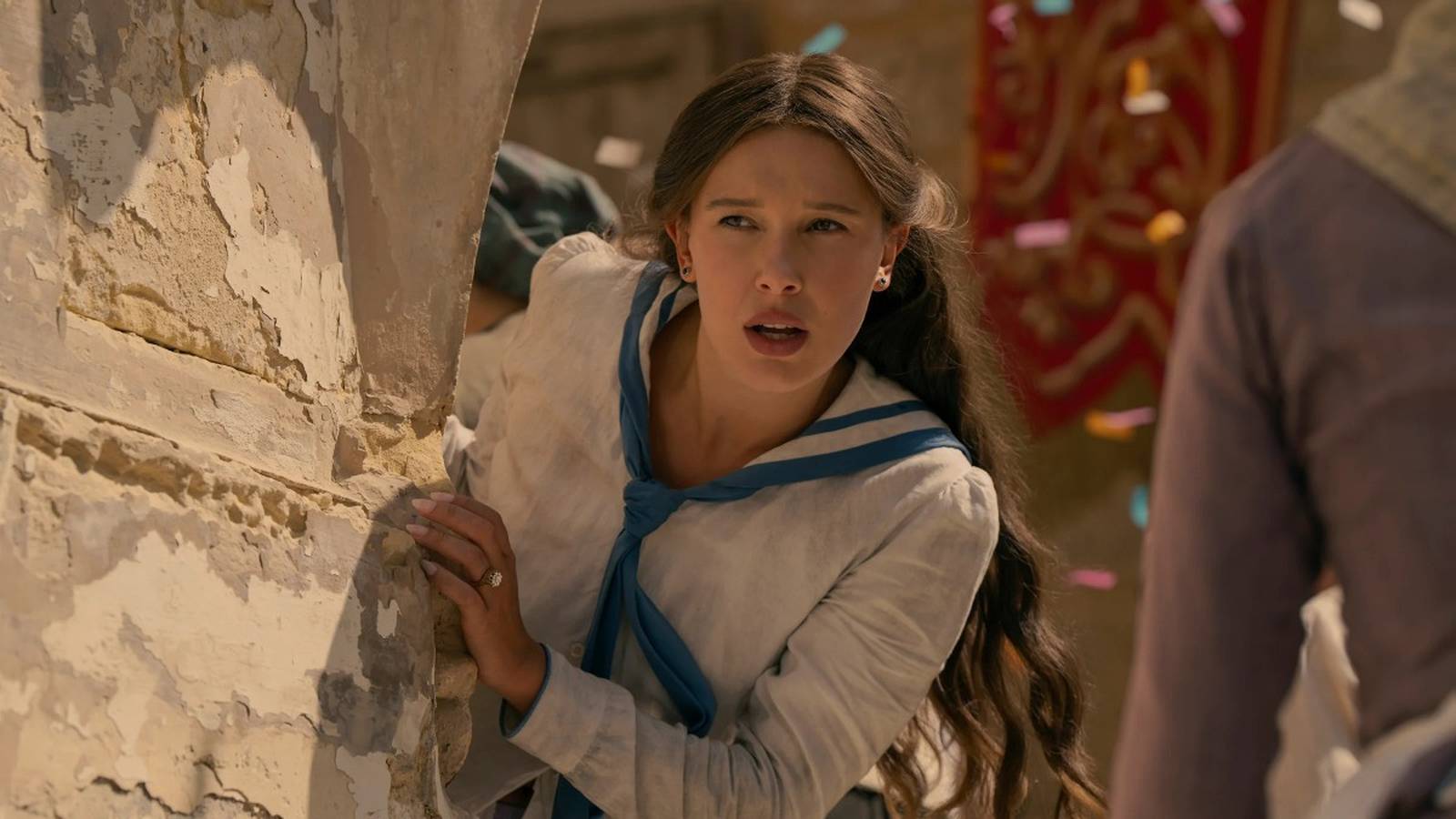 Millie Bobby Brown looking concerned while peeking around a stone wall in period costume