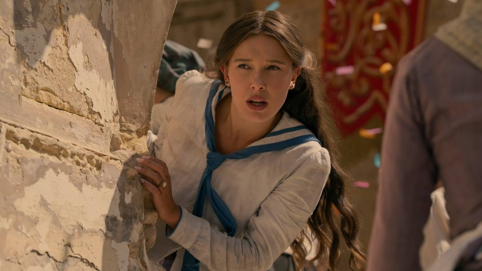 Millie Bobby Brown as Enola Holmes peeking around a stone wall in period clothing