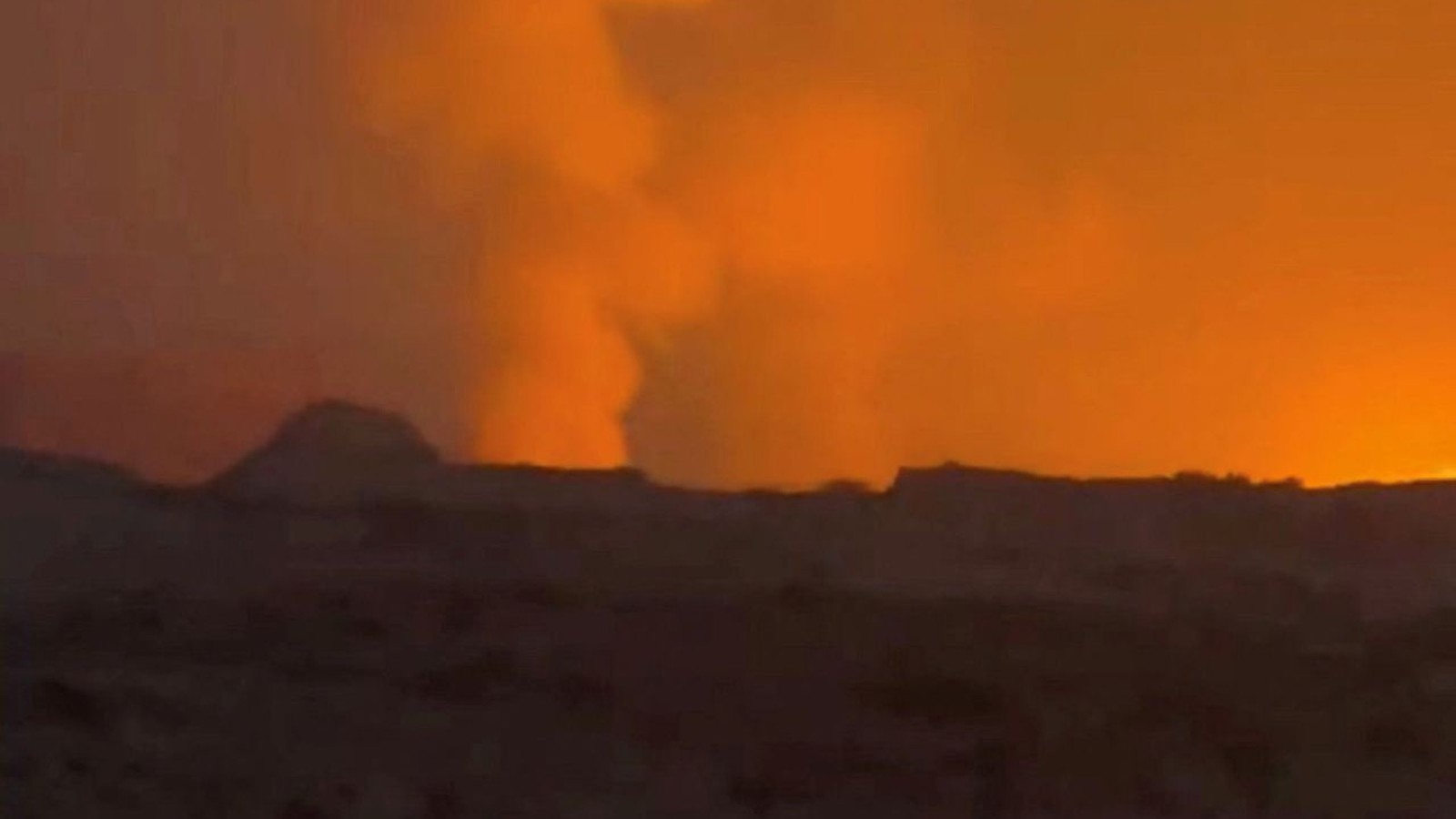 A large plume of smoke and fire rises from a desert landscape at night