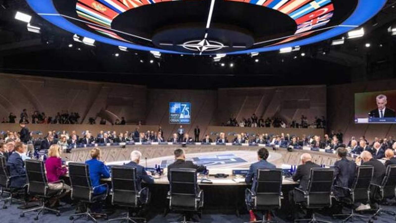 World leaders seated at a large circular table during a formal NATO summit meeting