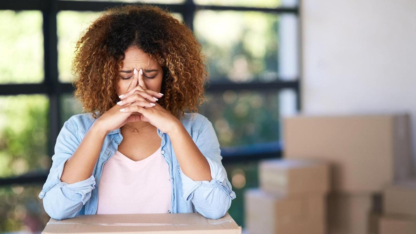 A stressed woman with curly hair resting her head on hands near moving boxes