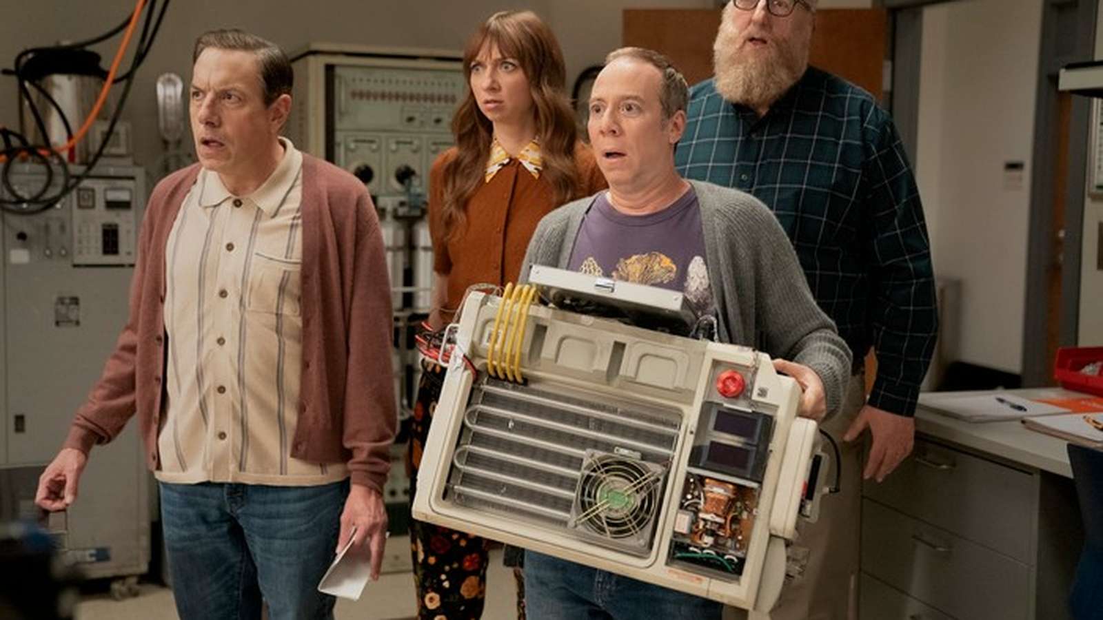 Four actors standing in a laboratory setting looking surprised while holding a device