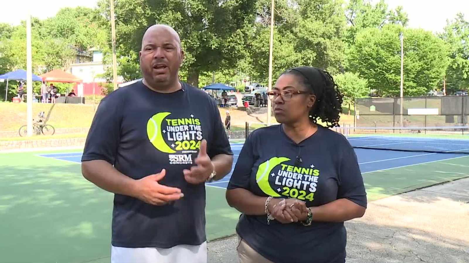 Two people wearing Tennis Under The Lights 2024 shirts standing on a tennis court