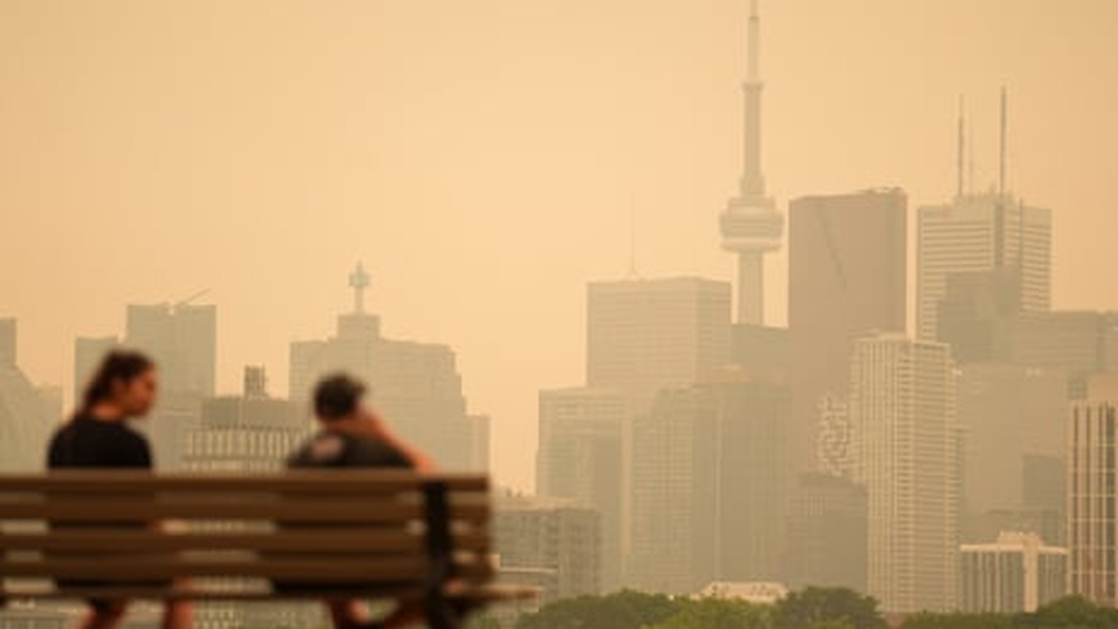 Two people sit on a park bench overlooking the hazy Toronto skyline