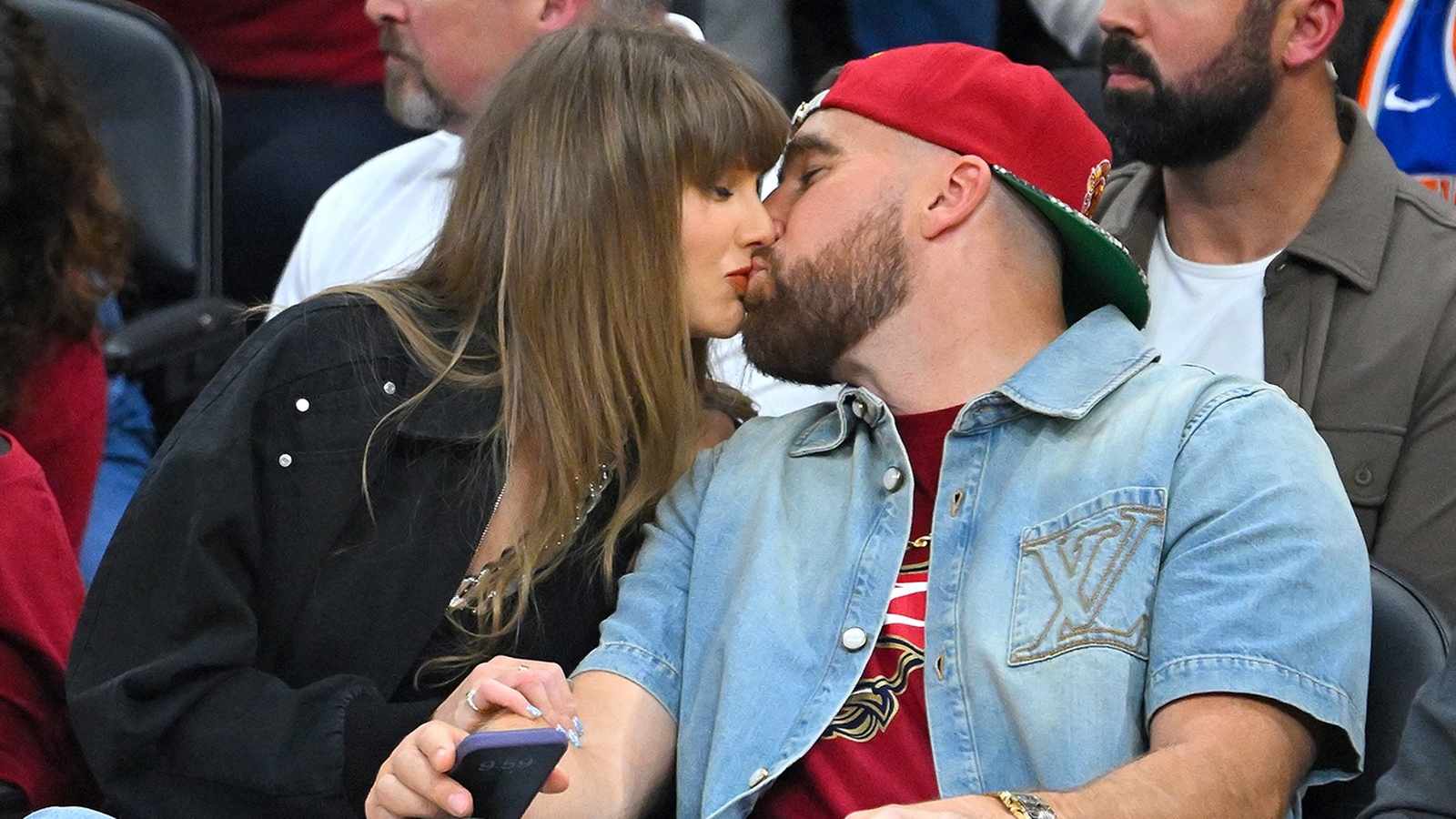 Travis Kelce and Taylor Swift sharing a kiss while sitting courtside at game