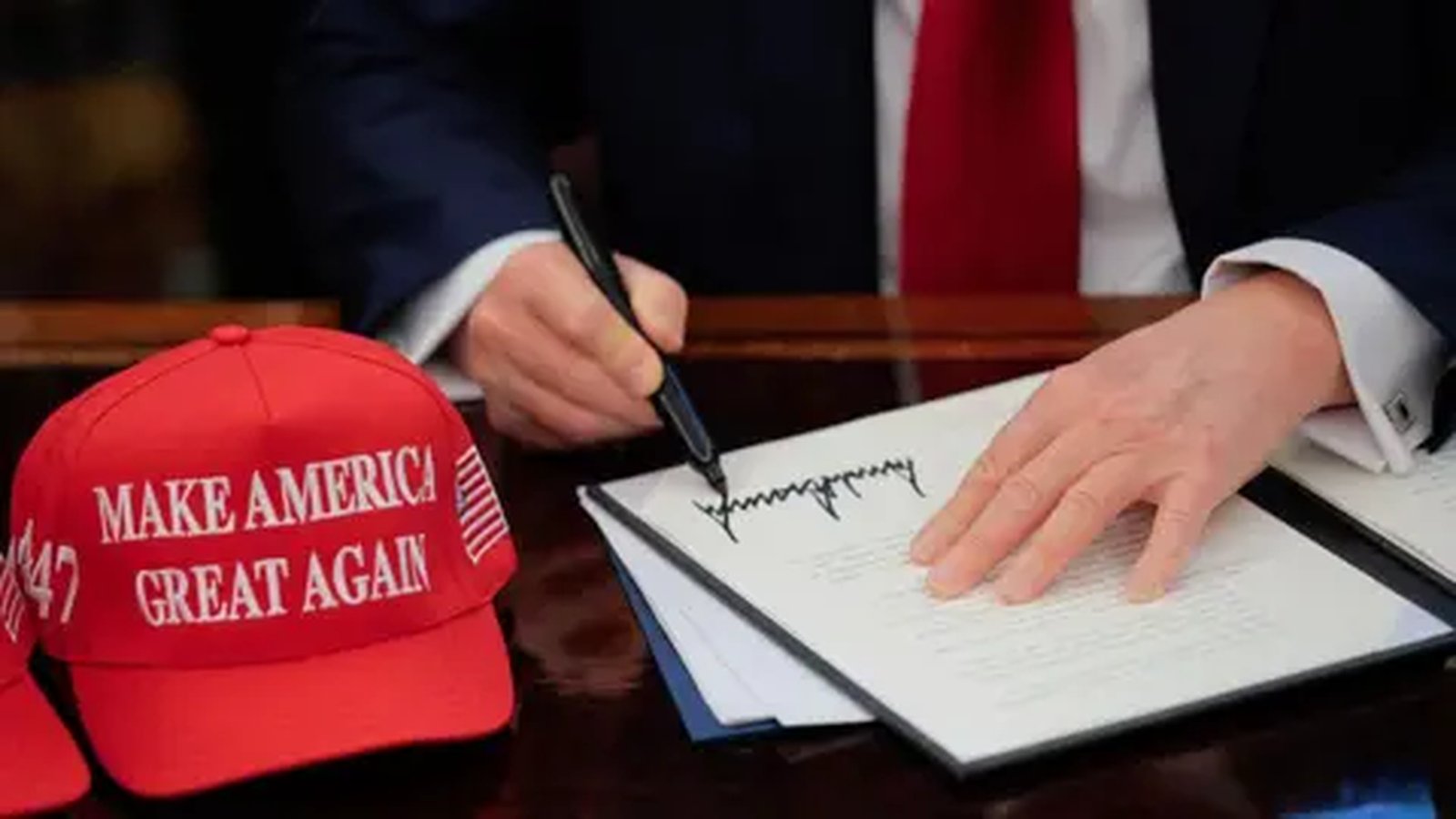 Donald Trump signing an official document with a red MAGA hat on the desk