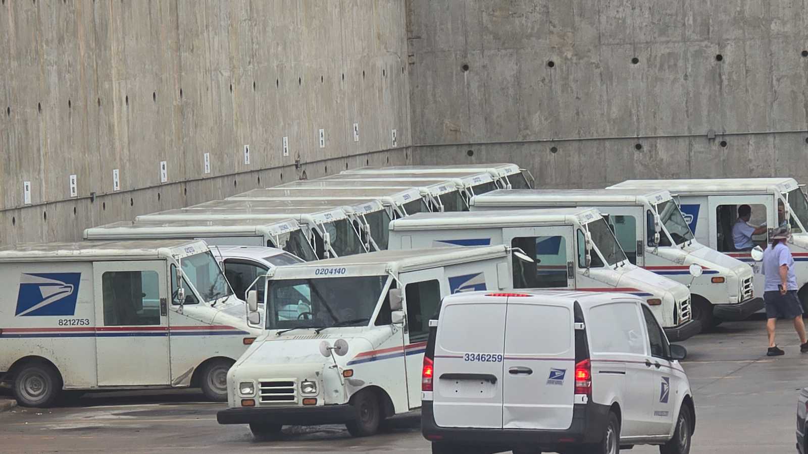 A row of white USPS mail delivery trucks parked in a concrete lot