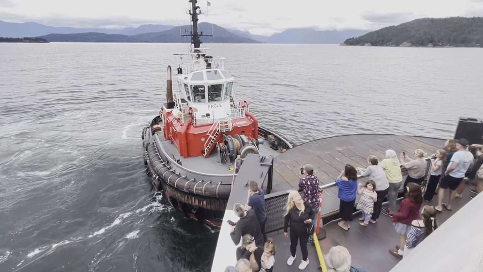 A red tugboat pulls a large BC Ferries vessel while passengers watch from the deck