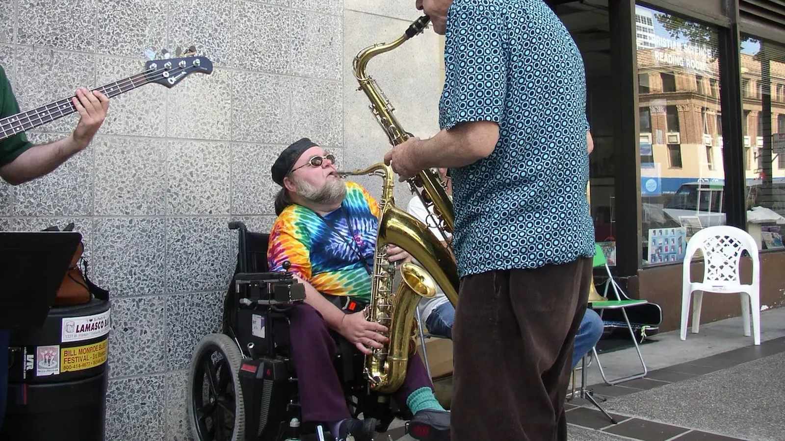 Saxophonist Bert Wilson in a wheelchair playing a saxophone alongside another musician outdoors