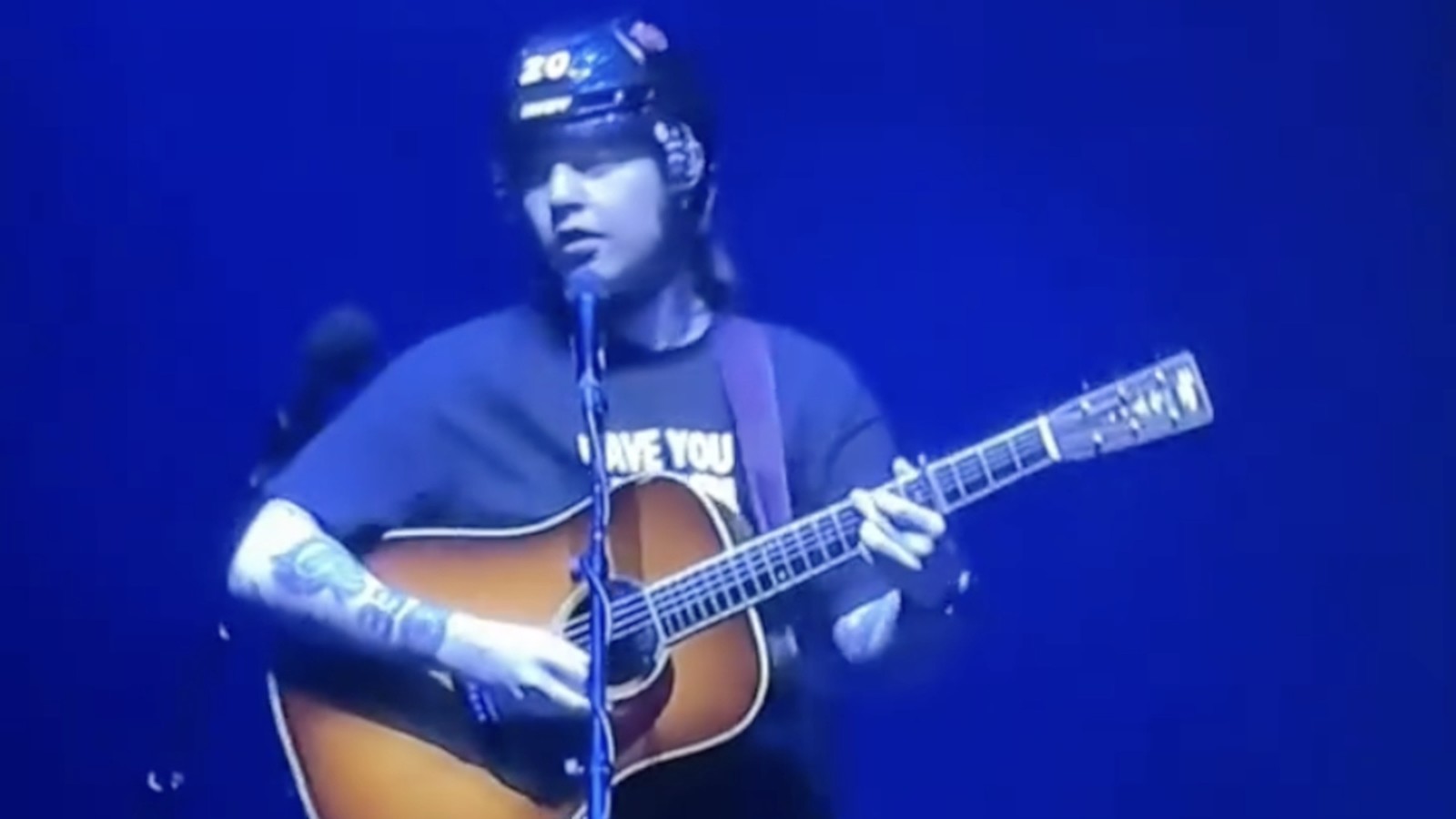 Billy Strings playing acoustic guitar on stage while wearing a protective hockey helmet