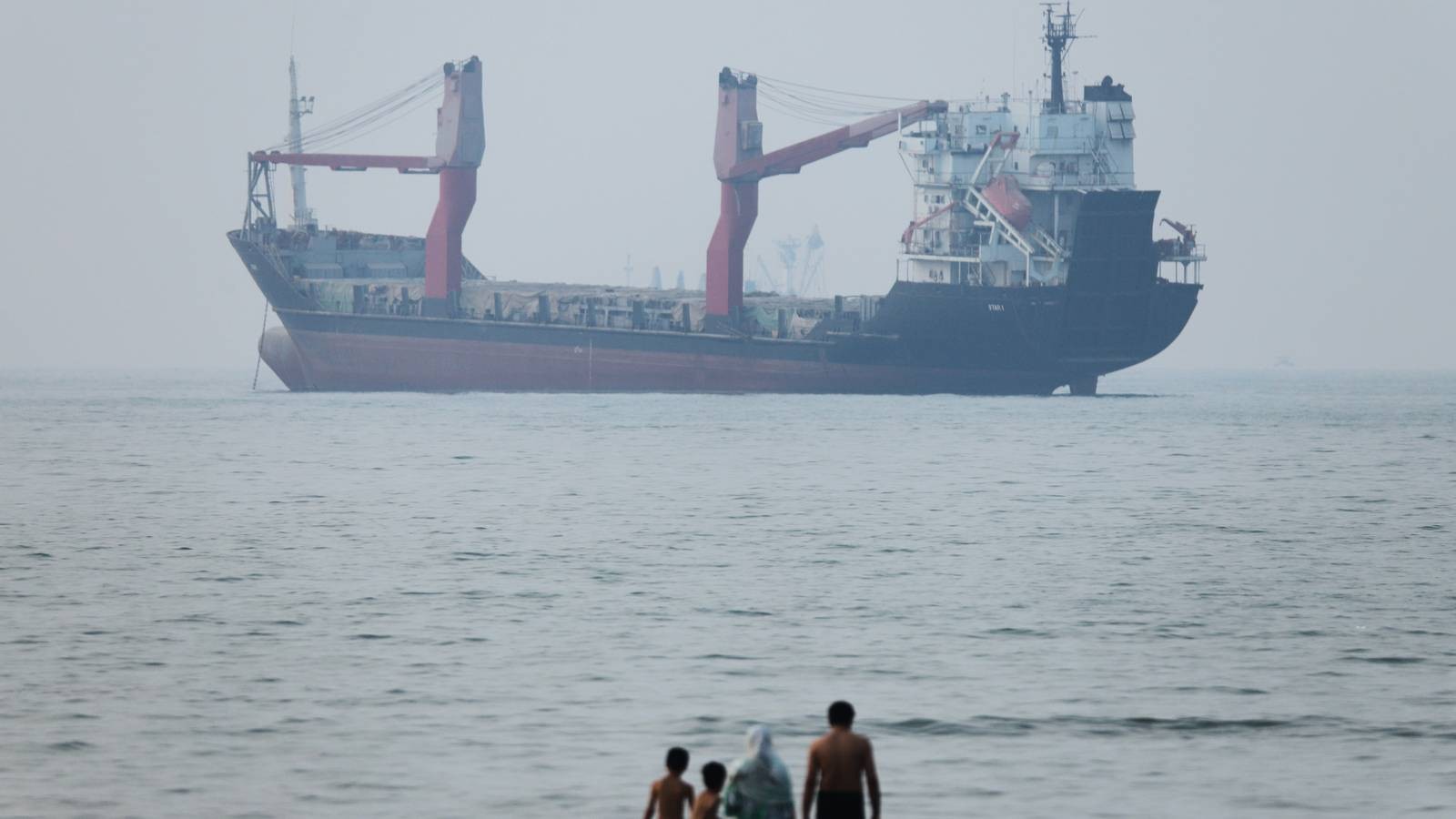 A large cargo ship sailing on the water with people standing in foreground