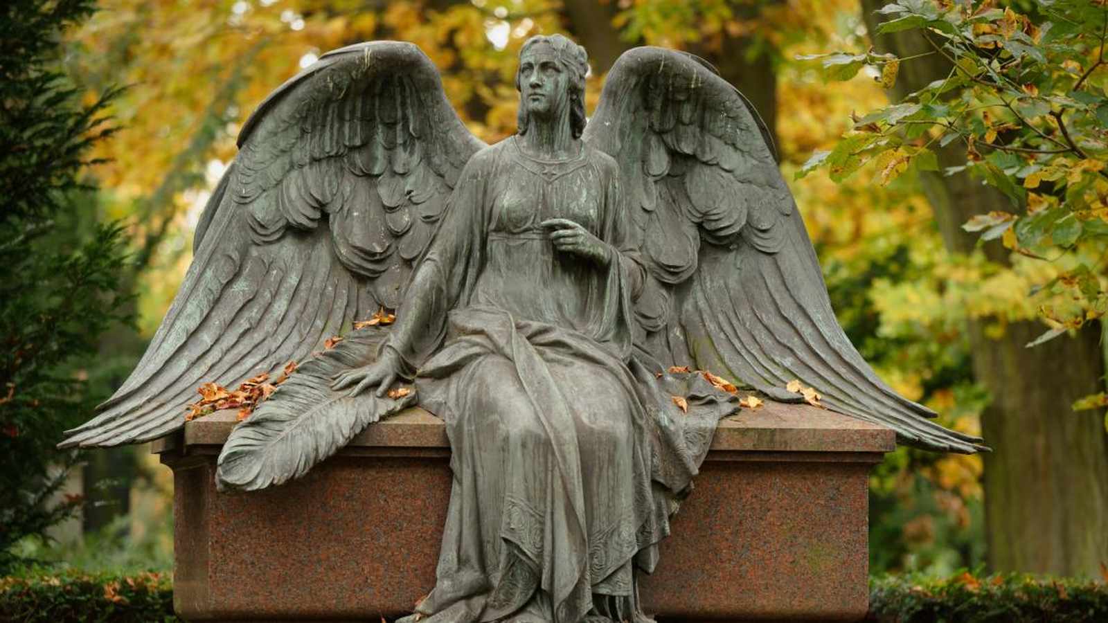 A bronze angel statue sitting on a stone tomb in an autumn cemetery