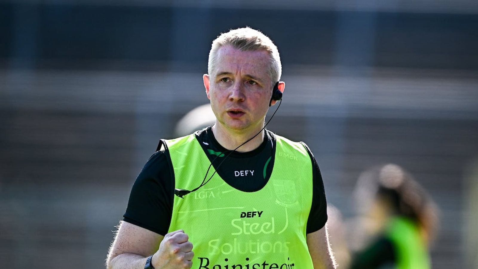 A GAA coach wearing a neon green bib standing on the pitch sideline