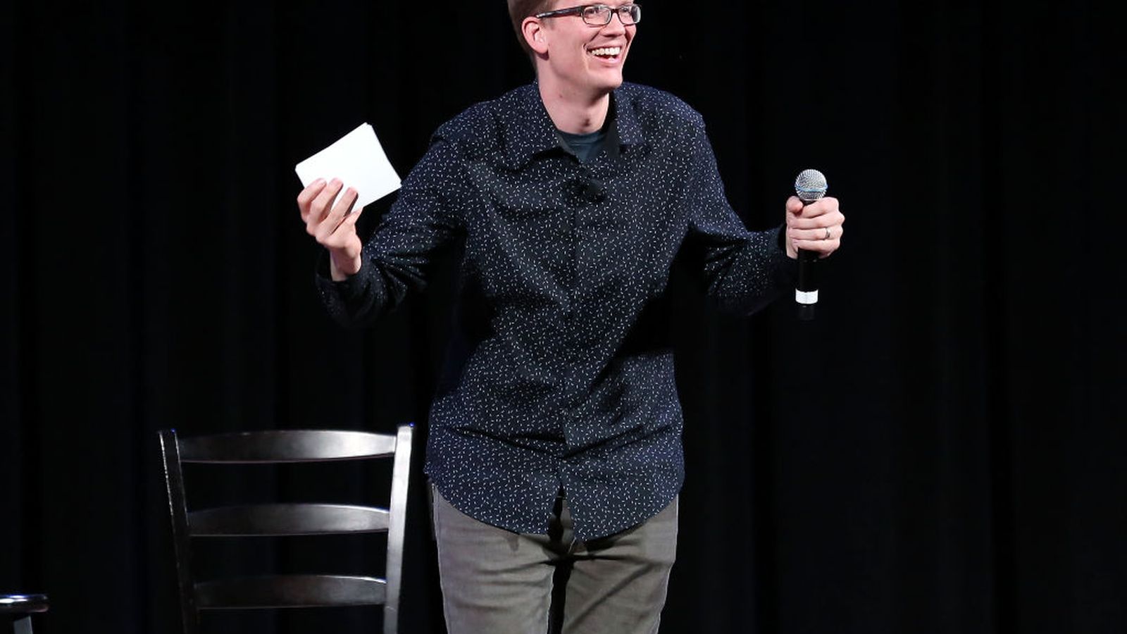 Hank Green wearing glasses and a patterned shirt holding a microphone on stage