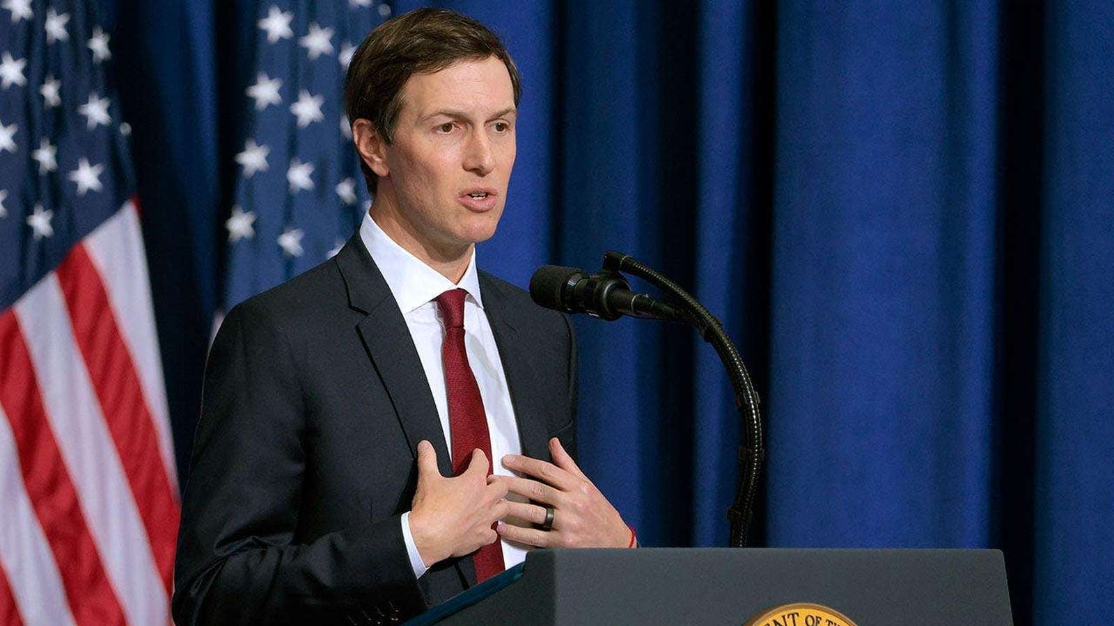 Jared Kushner wearing a suit and red tie speaking at a podium