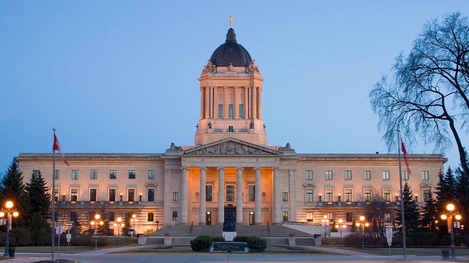 The Manitoba Legislative Building exterior illuminated at dusk with flags and surrounding trees