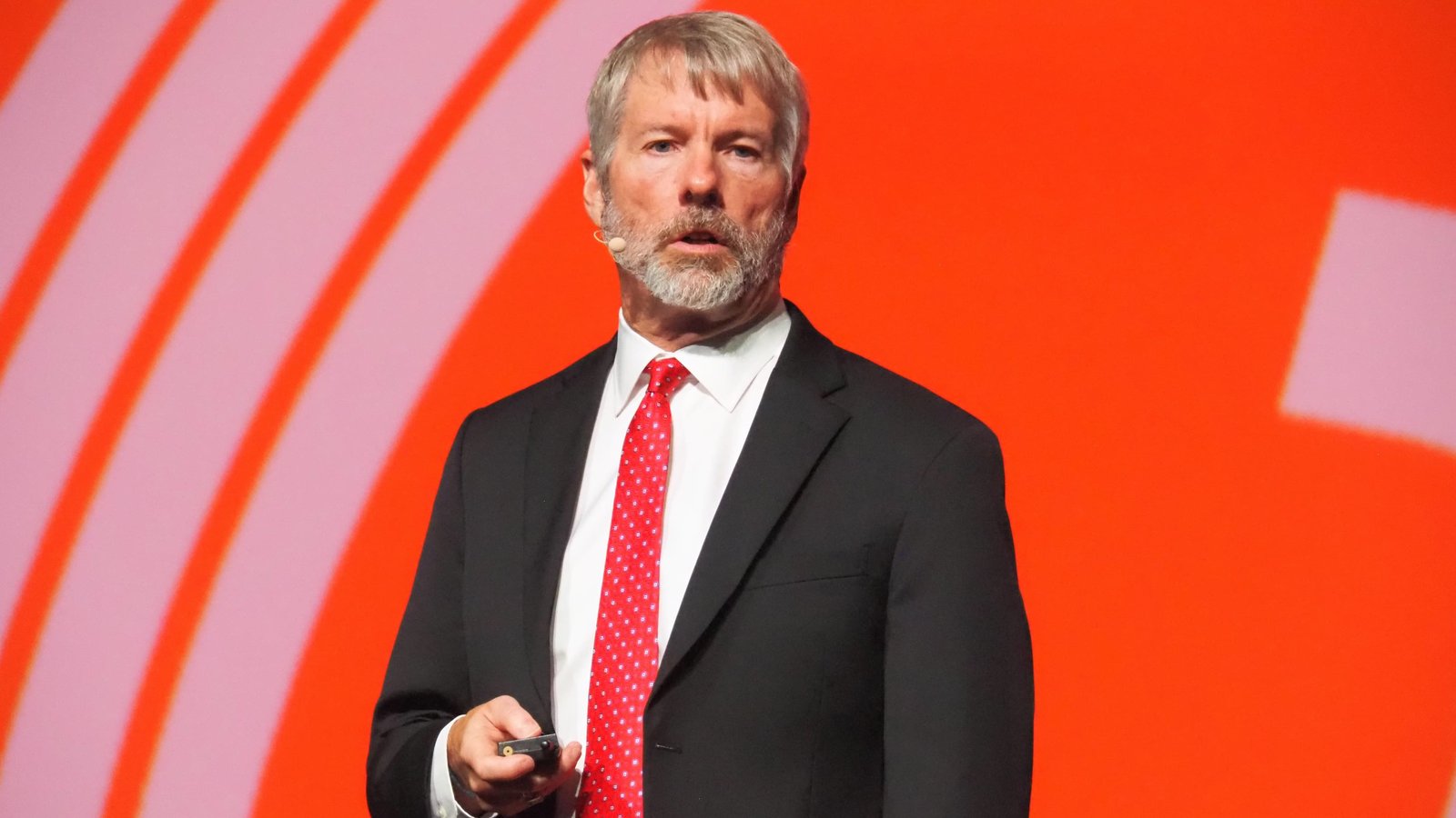 Michael Saylor wearing a suit and red tie speaking at a conference stage