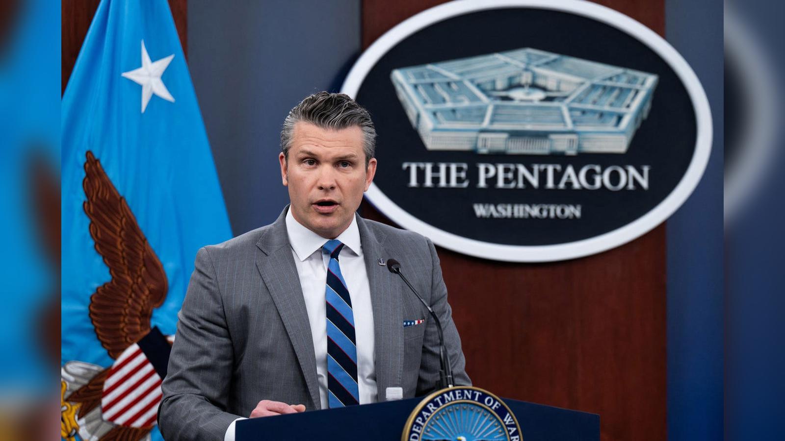 A male Pentagon spokesperson in a suit speaking at a podium with flags behind