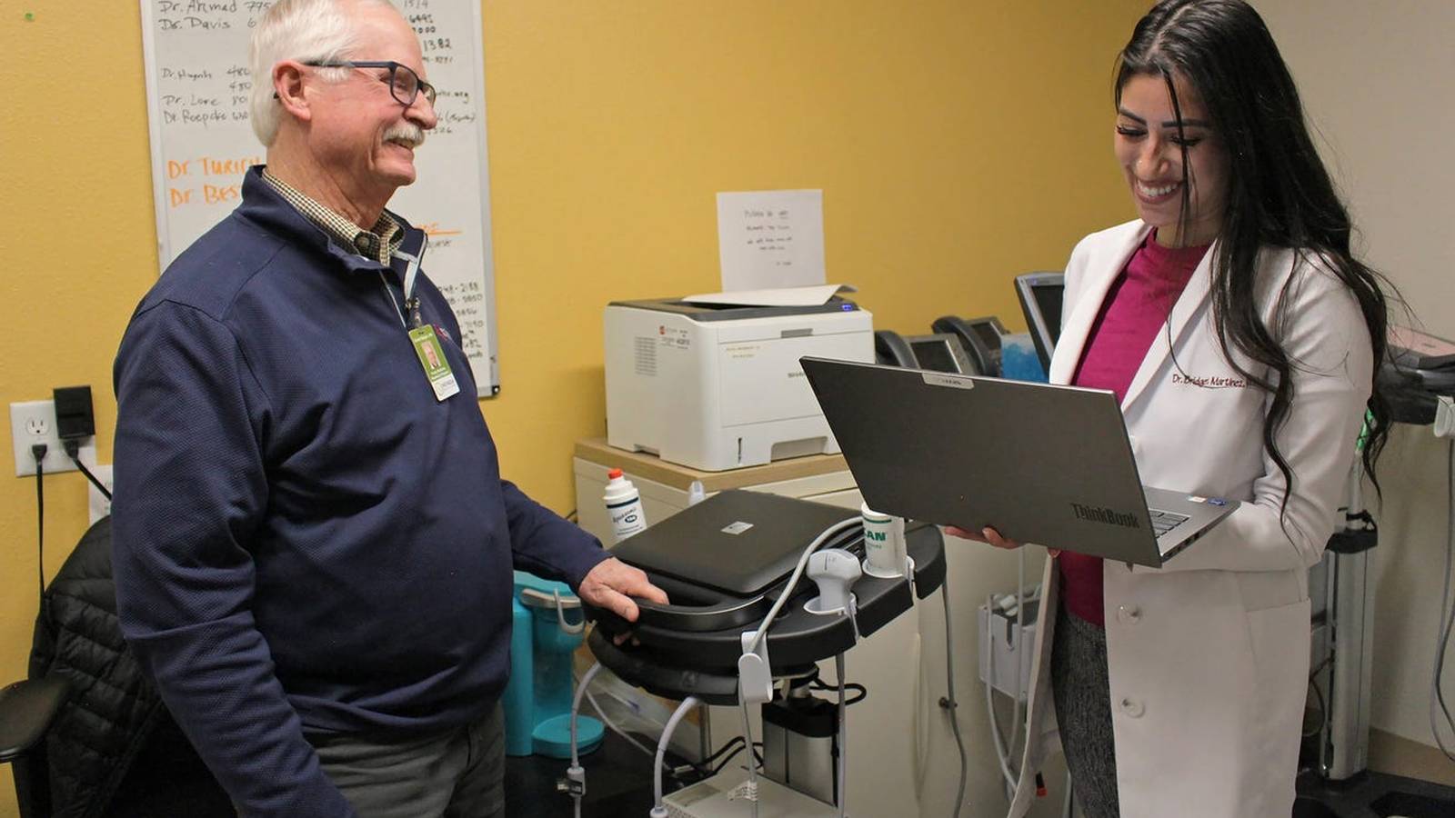 A female doctor in a white coat shows a laptop to a male patient