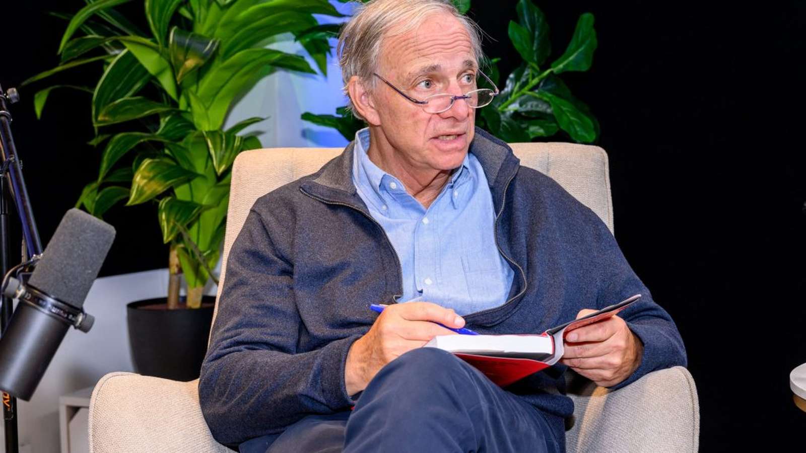 Ray Dalio sitting in a chair holding a book during an interview