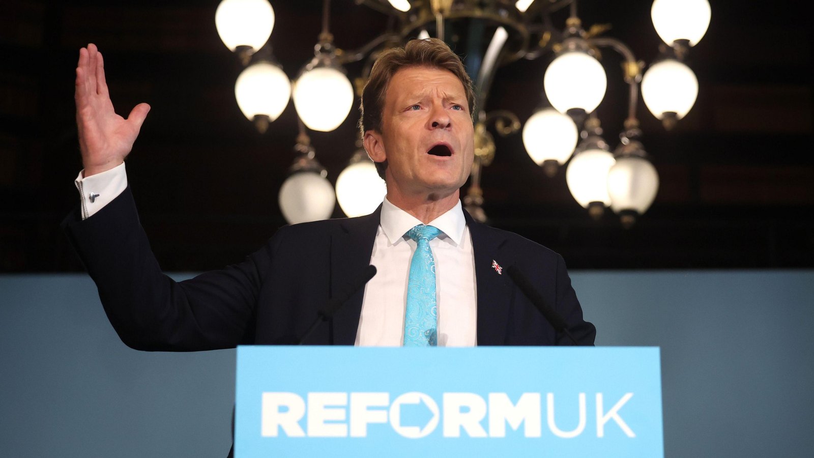 Richard Tice speaking at a podium with a Reform UK sign in front
