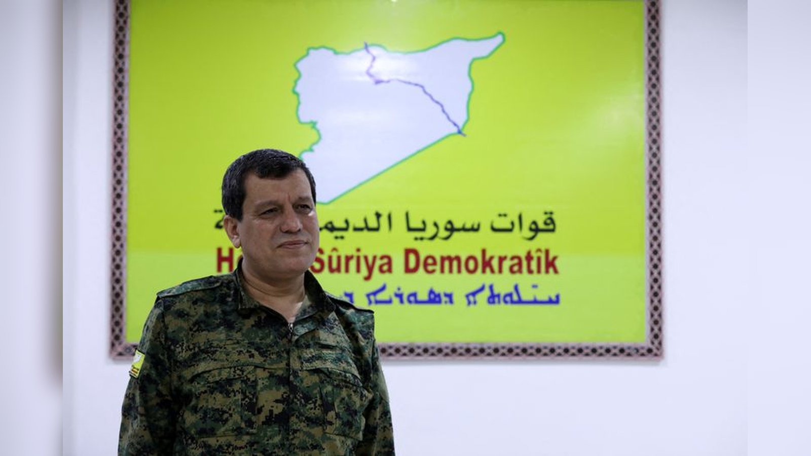 A Syrian Democratic Forces official standing in front of the official SDF organization banner