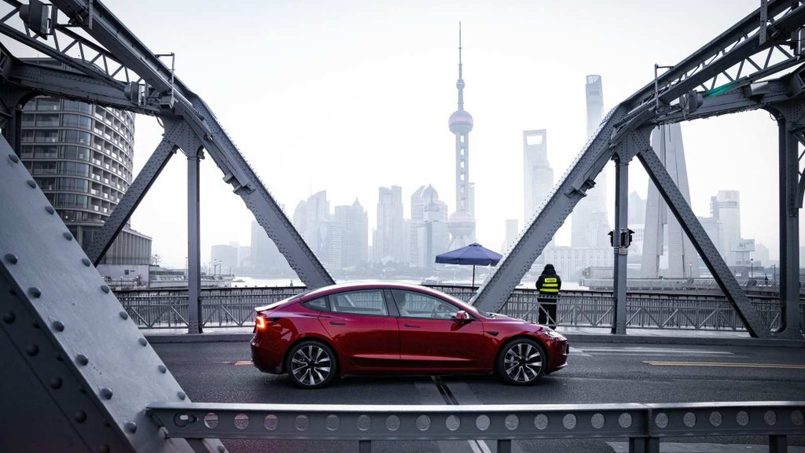 A red Tesla sedan driving on a bridge with the Shanghai skyline behind